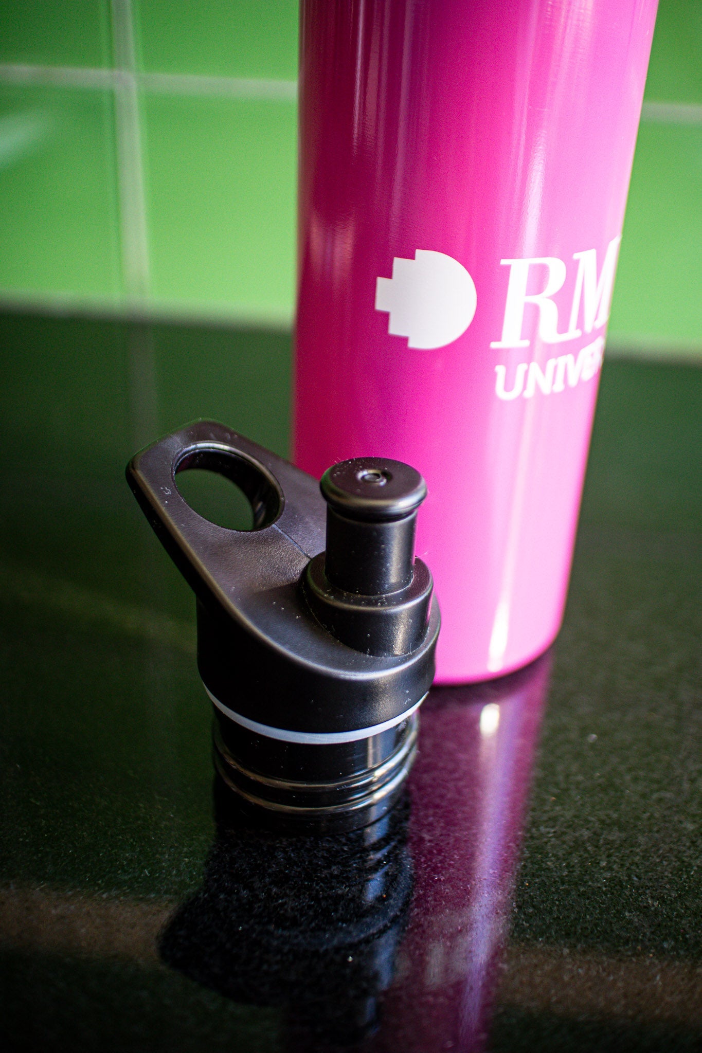 Pink RMIT bottle resting on a black tabletop with a green background, showing the left half of the white RMIT University logo printed on the bottle's body. The black cap is placed to the left side of the bottle. Available at the RMIT Store.